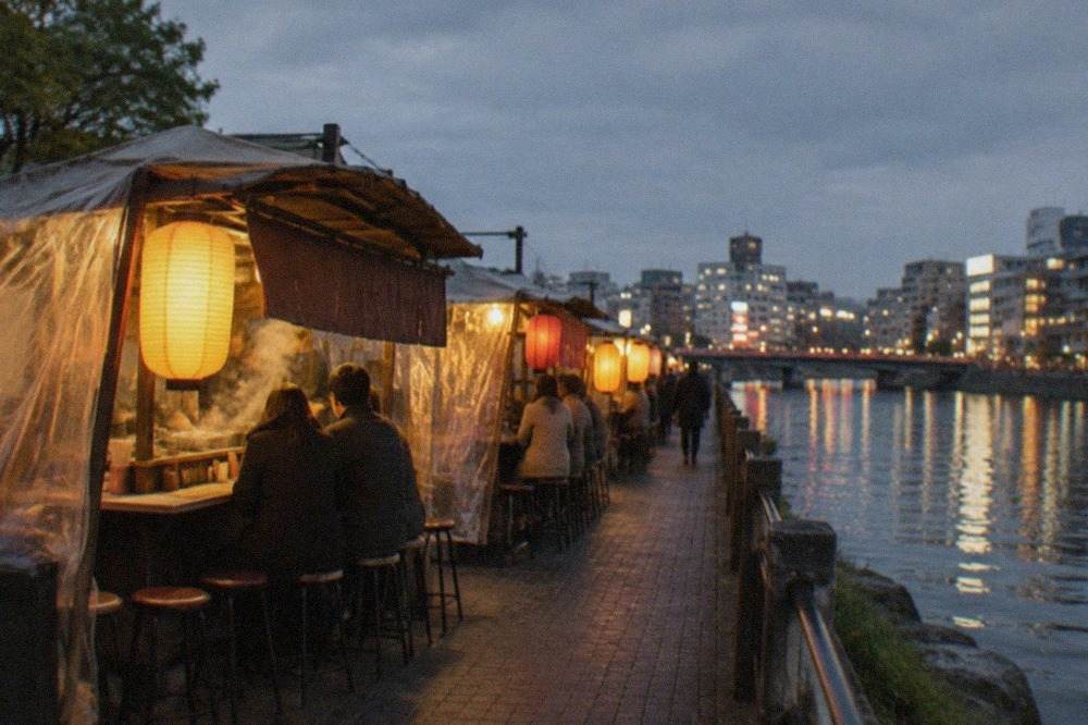 A row of glowing yatai food stalls along the Naka River at dusk, with city lights reflecting on the water.