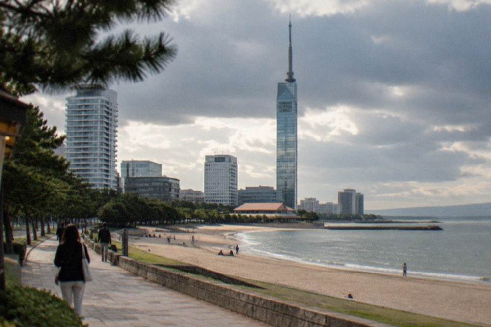Fukuoka Tower mirrored facade reflecting the sky, with Momochi beach and the Genkai Sea stretching behind it.