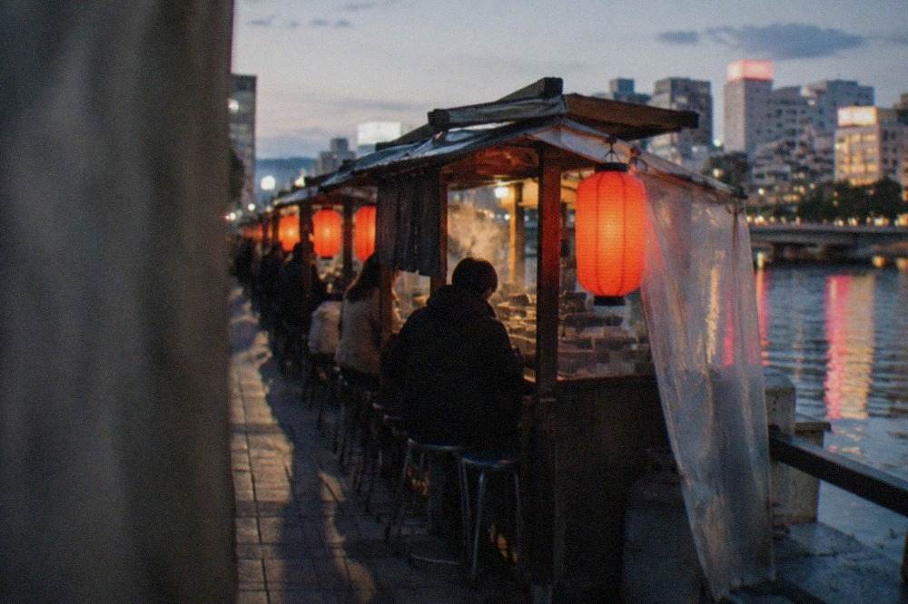 A row of glowing food stalls lined up along a river at night, with red lanterns reflecting on the water