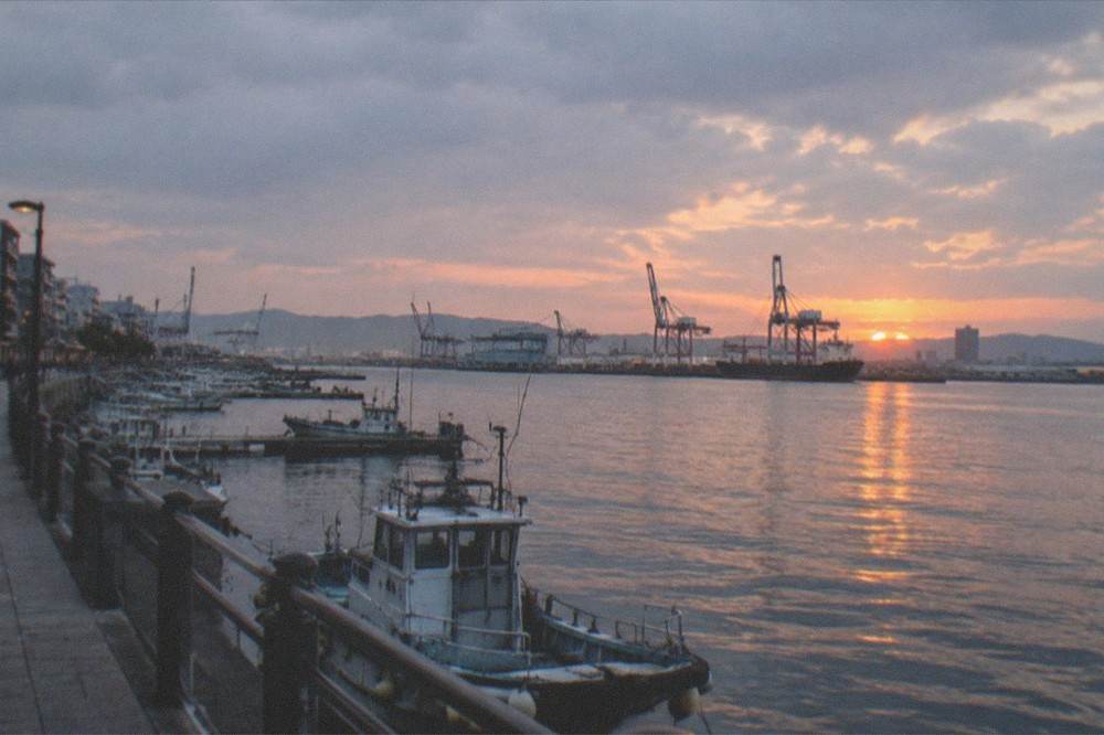 An evening view across Hakata Bay with port cranes and moored vessels silhouetted at sunset