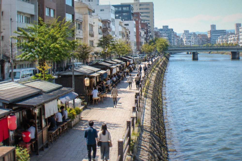 A riverside promenade with traditional wooden food stalls and modern buildings behind them