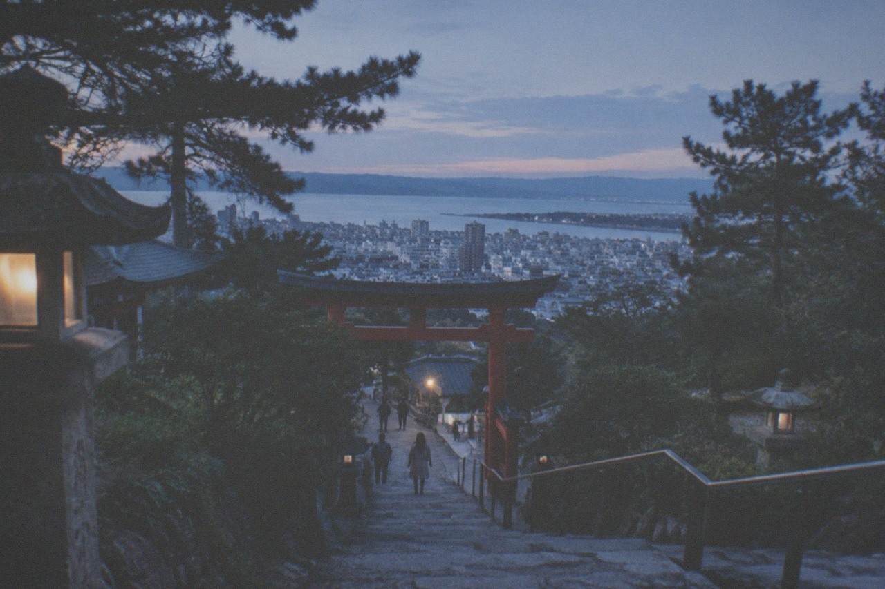 Stone steps rising through trees toward a vermilion shrine gate on a wooded hillside above the city