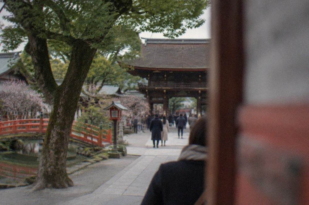Shrine forecourt with a large wooden gate and visitors standing under trees