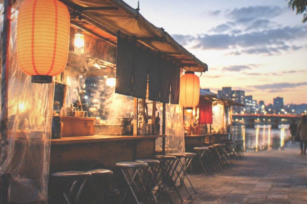 Food stalls glowing along a riverside path at dusk with bridge lights in the background