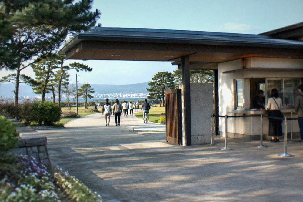 A park entrance gate and ticket window on a sunny morning, with trees and a wide path beyond