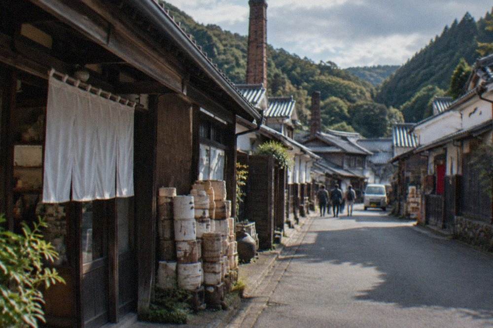 A quiet street of low porcelain workshops and kiln buildings beneath green hills in a small Japanese town