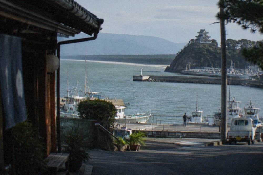 A Japanese castle keep on a hill above a wide bay, with pine-covered sandbar curving along the shoreline in the distance