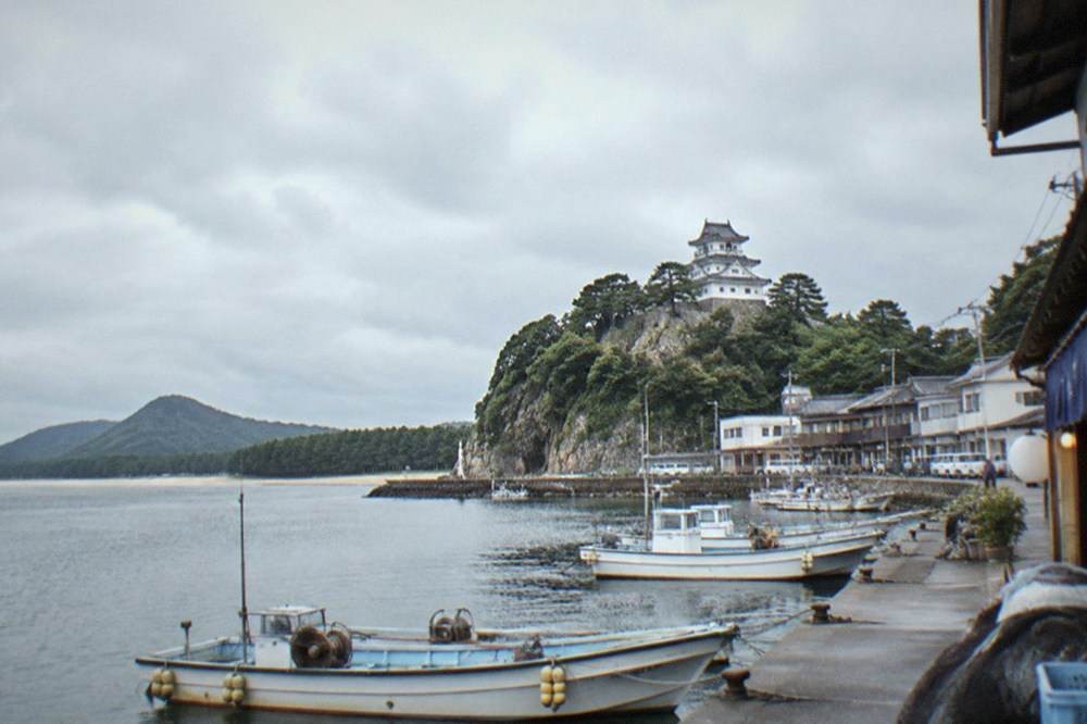 A white-walled castle keep on a headland, seen from a fishing harbour with small boats moored below