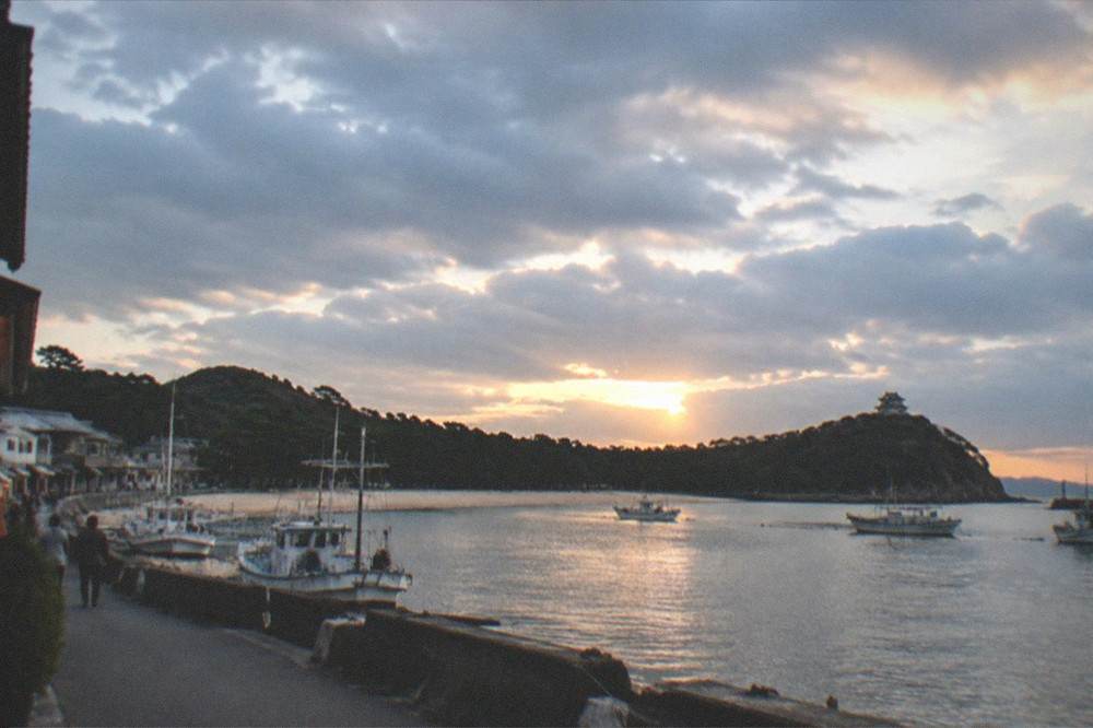 A calm bay at dusk with fishing boats at anchor and a castle silhouette on the headland
