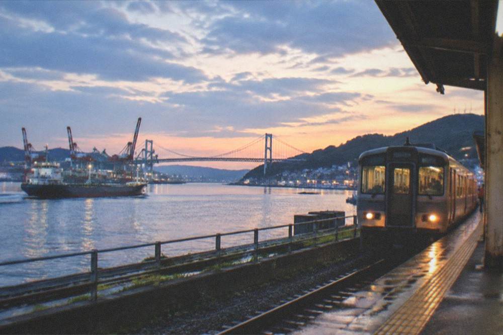 Railway platform at dusk with a local train at the platform and city lights beyond