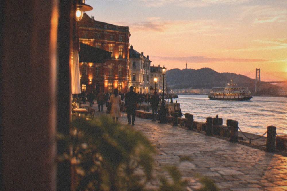 A waterfront promenade at dusk with lit heritage buildings and a ferry crossing the strait