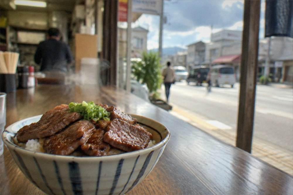 A steaming bowl of rice topped with sliced grilled beef served on a wooden counter
