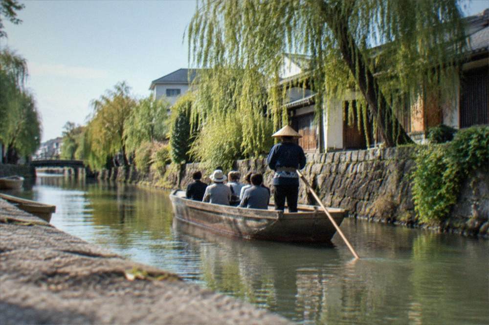 A punt boat passing a weeping willow and stone canal wall in soft late afternoon light