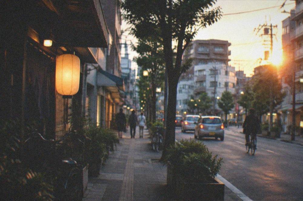 A city street at dusk with illuminated shop signs and a lit cafe window