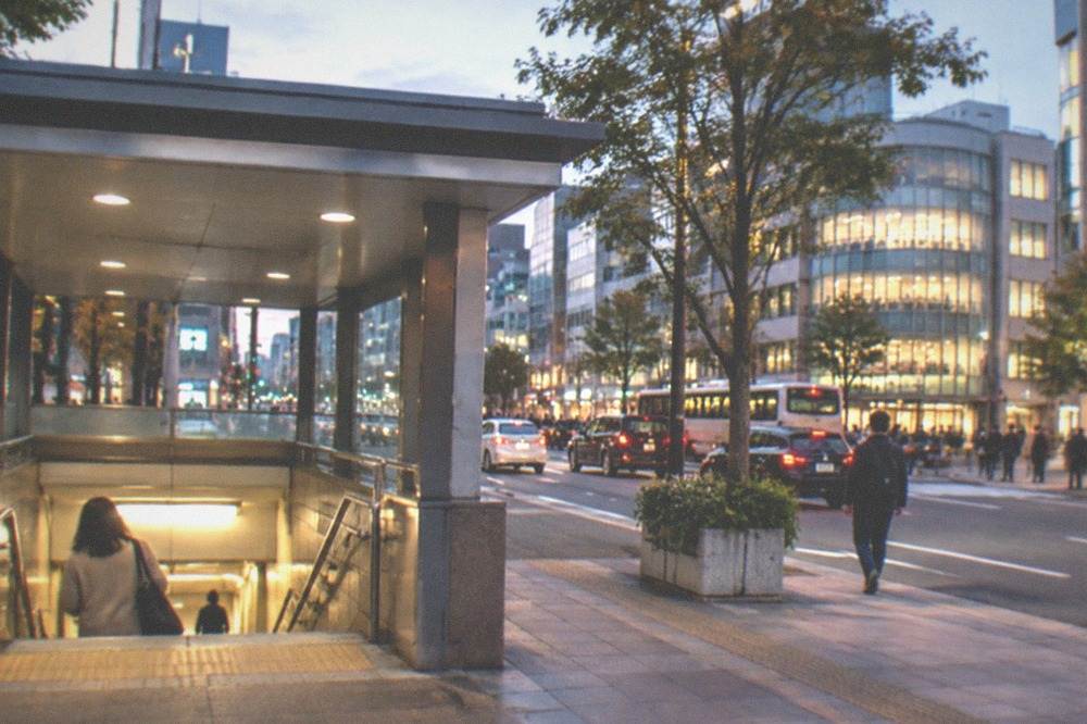 A busy city intersection at dusk with illuminated buildings and a subway station entrance on the corner
