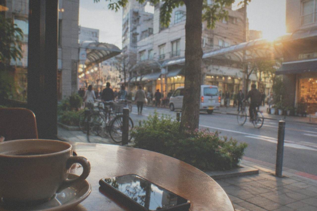 A smartphone resting on a cafe table beside a cup of coffee, with a city street and illuminated shopfronts visible through the window