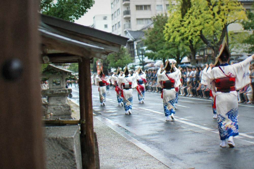 Costumed parade participants dancing down a wide city avenue lined with spectators