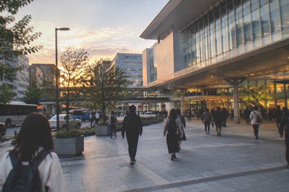 A wide view of Hakata Station's exterior at dusk with travellers walking past with small day packs