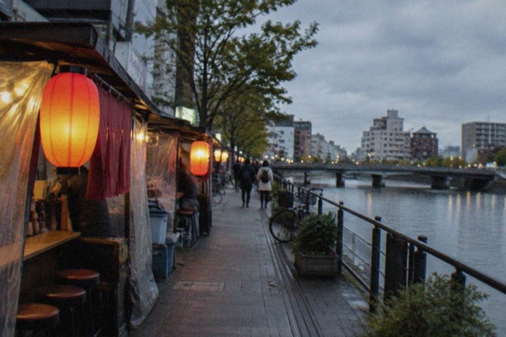 Food stalls with lanterns glowing along a riverside street in the evening