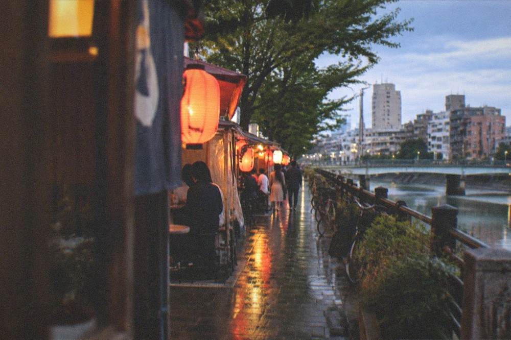 A row of illuminated food stalls with lanterns glowing along a riverside street at night