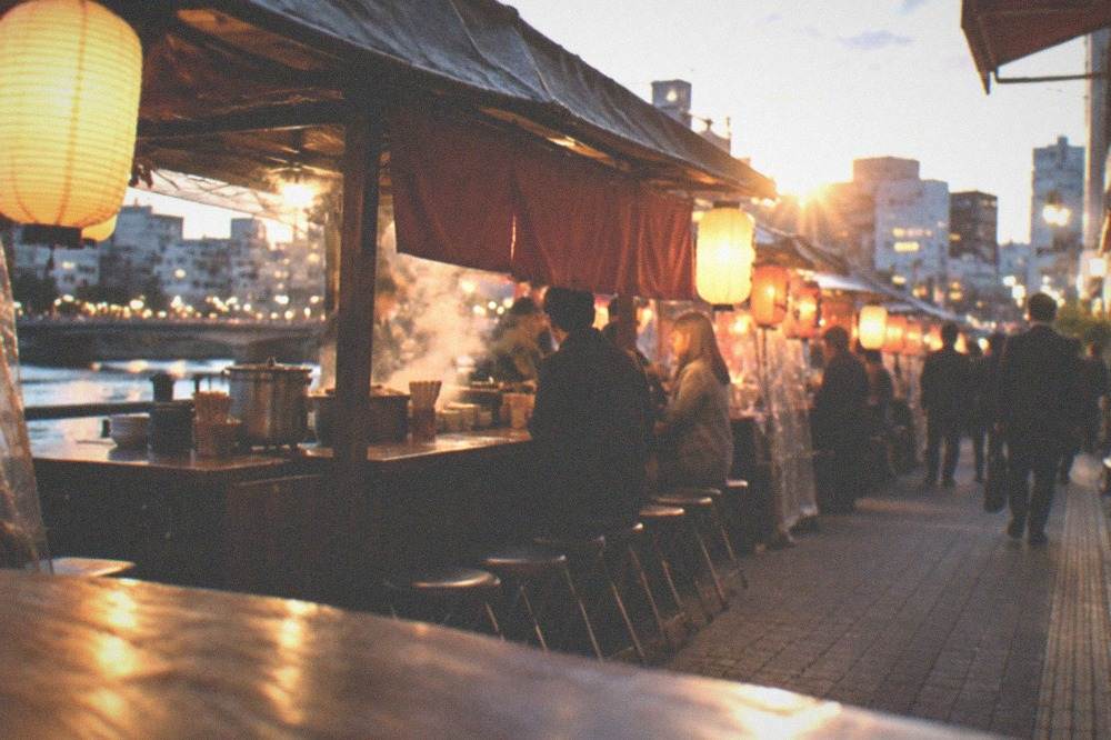 A narrow city street at night with food stall awnings, steam and office workers queueing