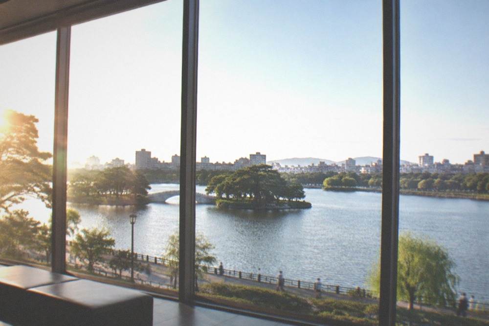 Tall windows of a museum gallery overlooking the Ohori Park lake and tree line
