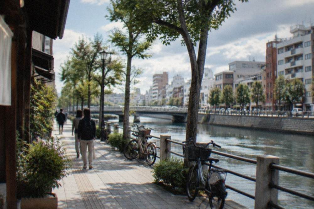 A compact Japanese city street with a canal, modern buildings and pedestrians on a wide sidewalk in daylight