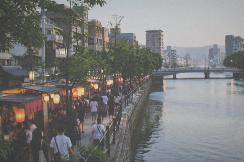 Crowds and food stalls along a river promenade at night with lanterns glowing