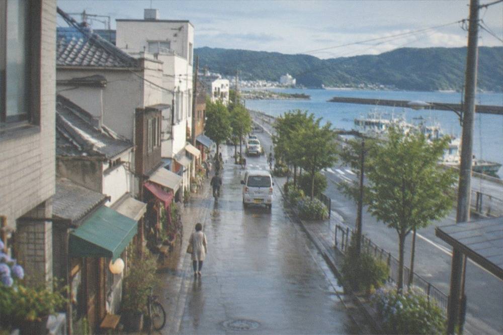 A coastal road along a calm blue bay with small fishing boats moored at a harbour and green hills behind.