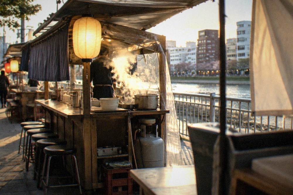 Steam rising from a small food cart kitchen while a cook prepares noodles under a glowing lantern