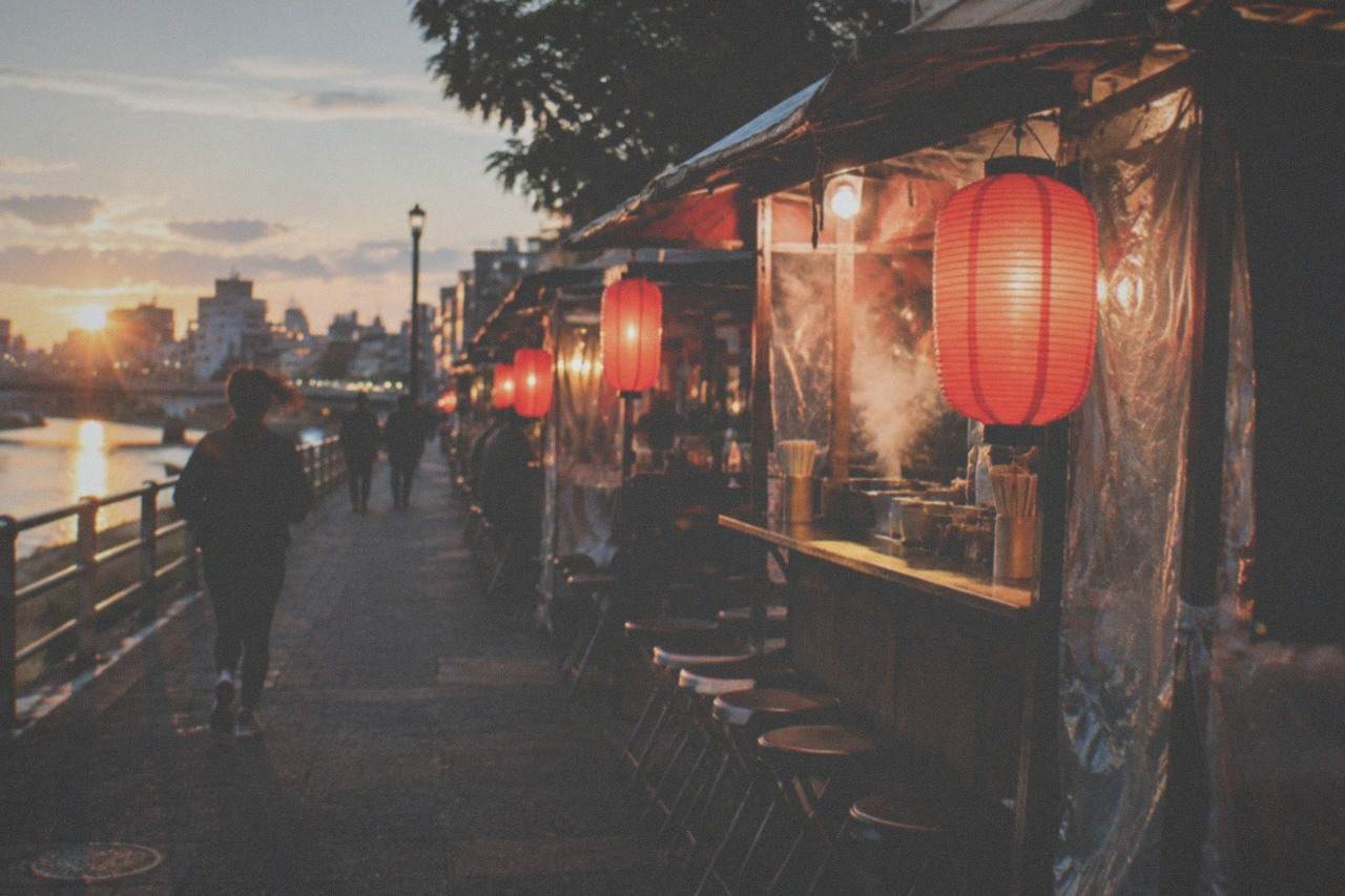 Row of small lit food stalls with red lanterns lining a riverside street at night
