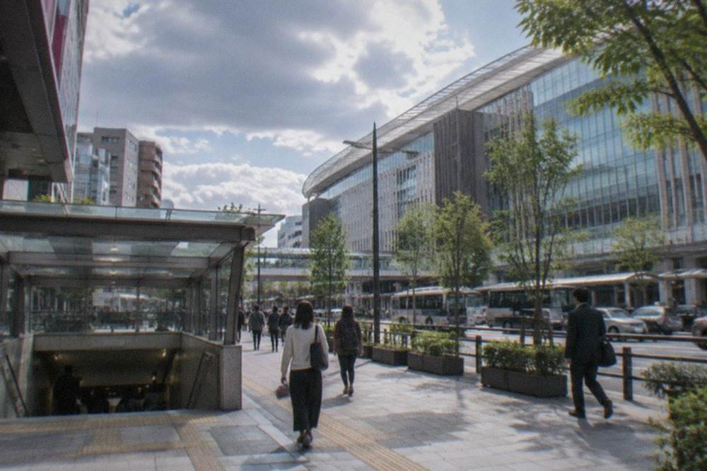 A Fukuoka city street with a subway station entrance and pedestrians on a wide sidewalk