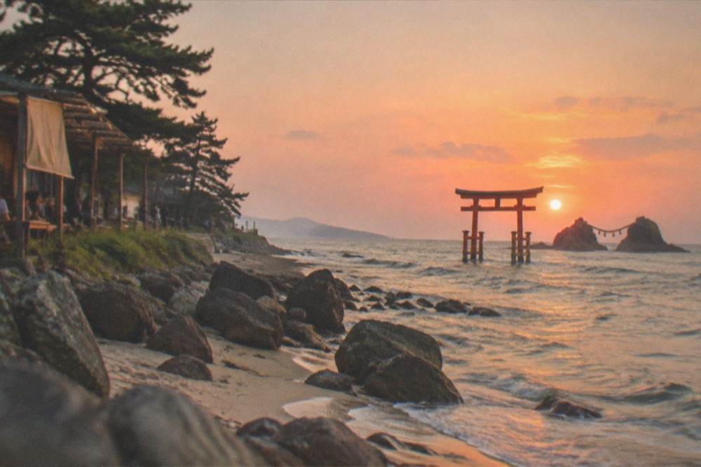 A red torii gate and paired rocks standing in shallow sea water under an orange evening sky