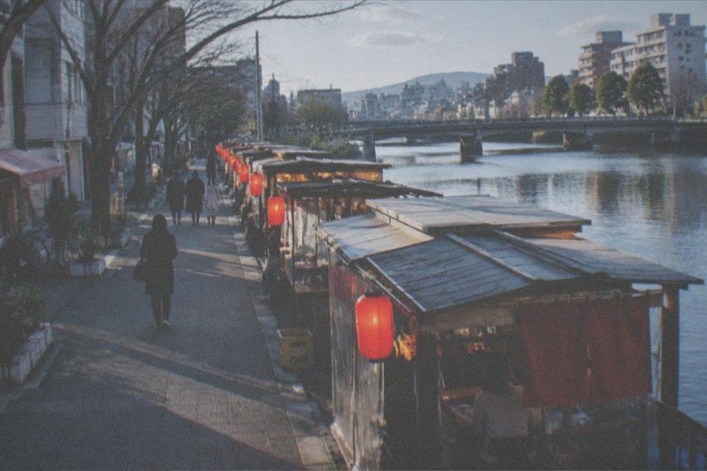 Small lit food stalls lined up beside a river at night with red lanterns