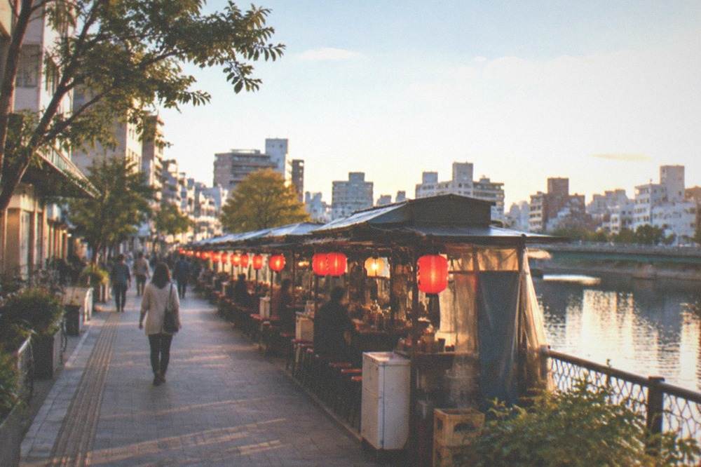Row of small lit food stalls along a river at night with red lanterns reflecting on the water