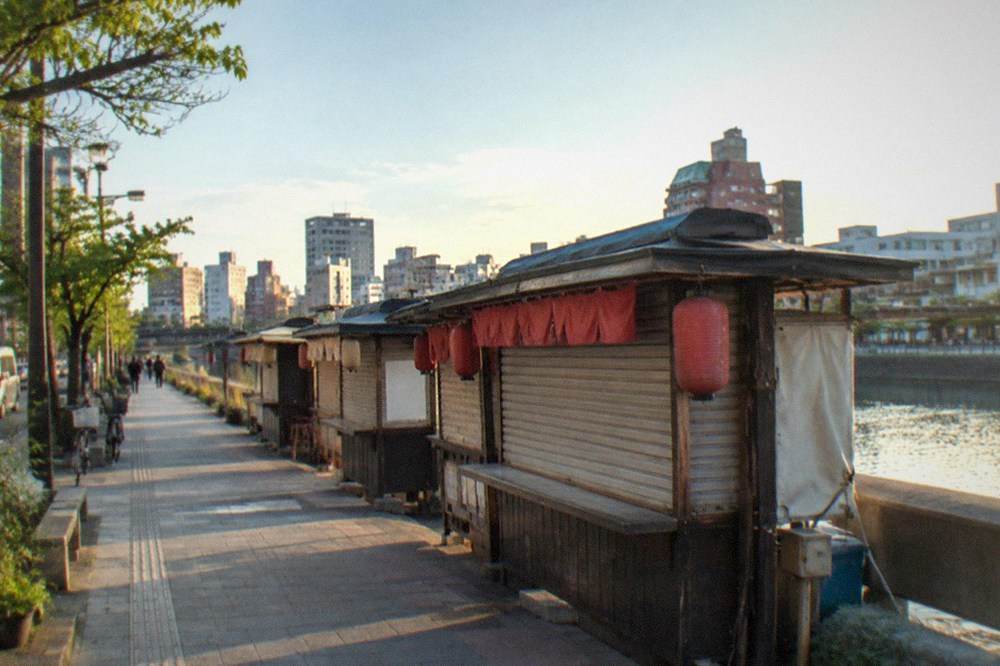 Food stalls lined up along a river at dusk with lanterns unlit and shutters closed