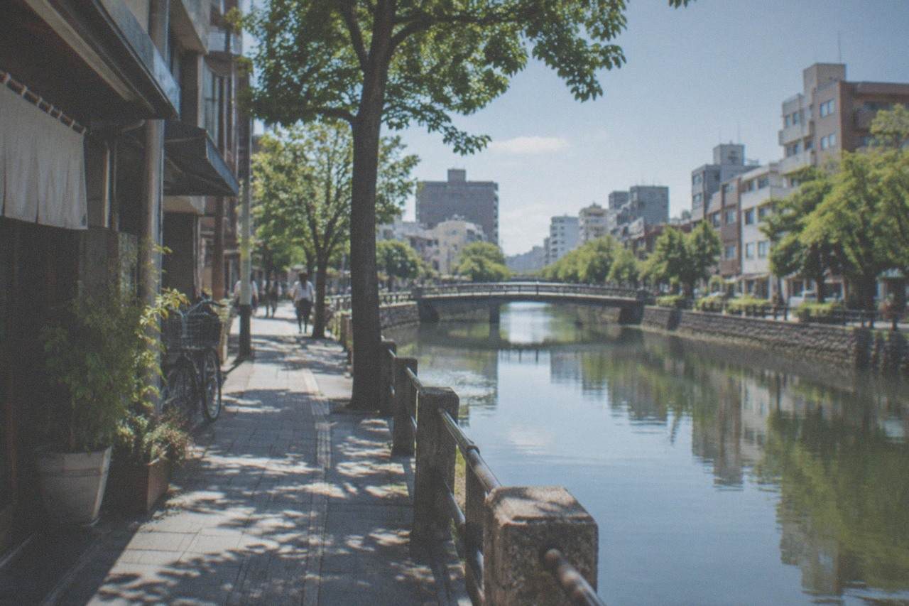 A quiet canal-side path in Fukuoka with trees reflected in the water on a clear morning