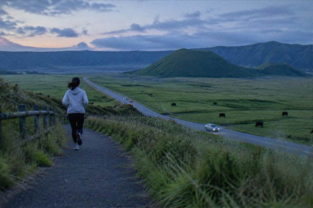 Grassland slopes around the Mount Aso caldera under drifting clouds with a winding road below
